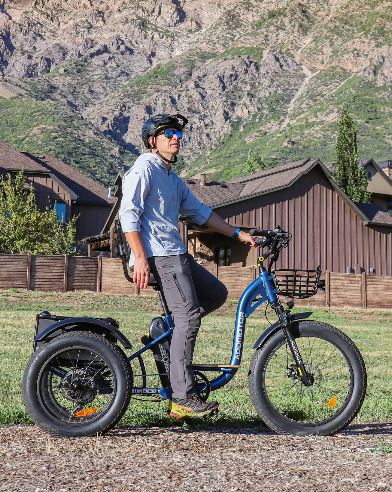 A man sitting on Grandtan fat tire electric tricycle with a blue frame and large tires, designed for off-road use. The tricycle is equipped with a front basket, and the backdrop features mountains and houses, highlighting its versatility and suitability for outdoor adventures.