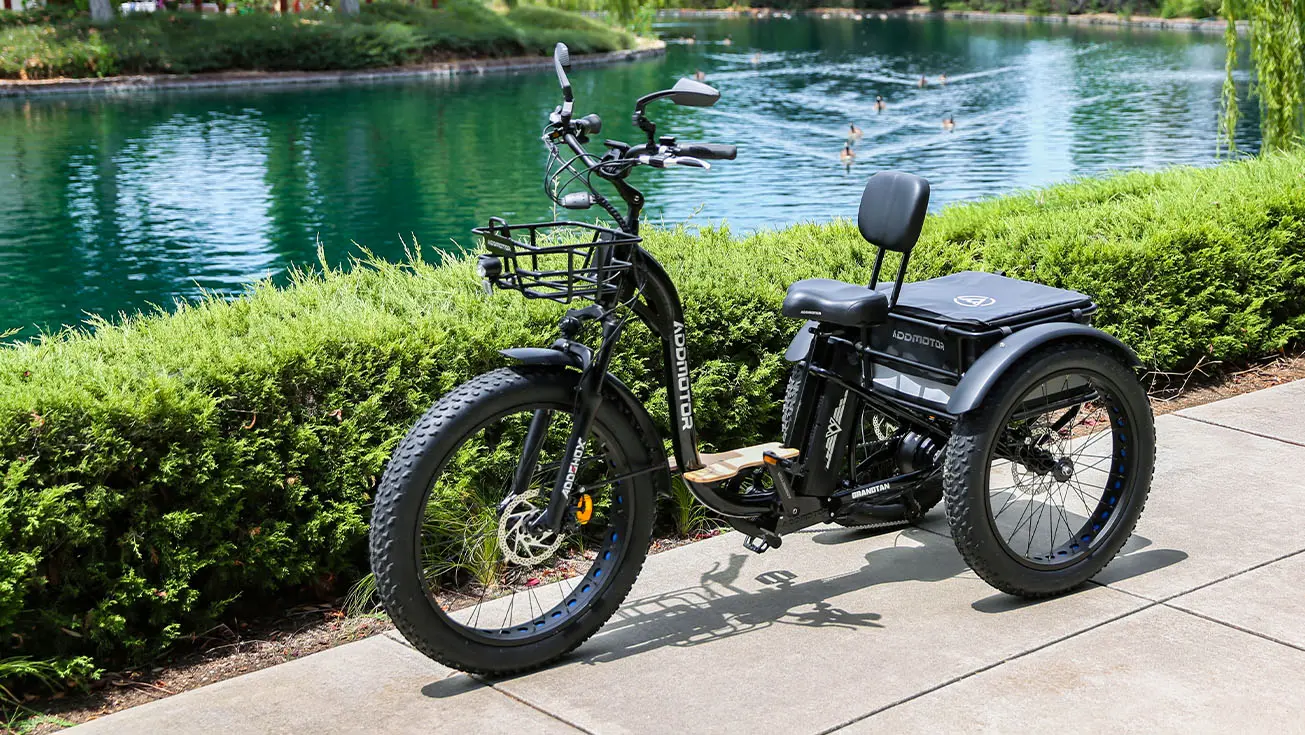 Grandtan M-340 trike parked on a sidewalk next to a scenic lake, featuring robust fat tires, a sturdy frame, and a spacious rear cargo area. The trike is ideal for off-road adventures and offers stability and comfort with its ergonomic padded seat and handlebars.