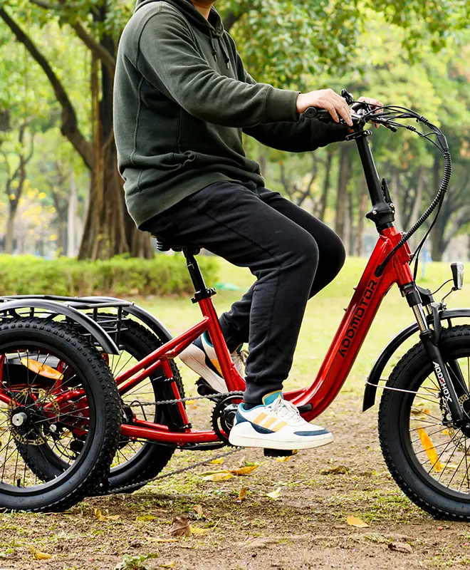 a man wearing a green coat and sport shoe is riding a red addmotor folding trike on the mud in a garden with a lot of trees
