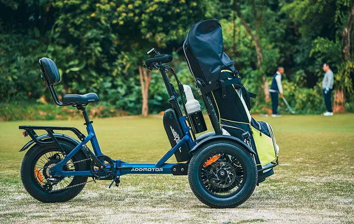 Reverse trike designed for golf with two front wheels, a rear wheel, and a storage area for golf bags, positioned on a grassy golf course.
