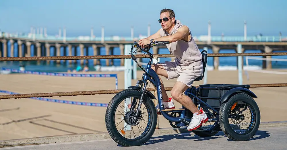 E-bike trike ridden along a beachfront promenade, featuring a sturdy frame, three wheels, and a rear cargo area, set against a sunny coastal backdrop with volleyball nets and a pier.