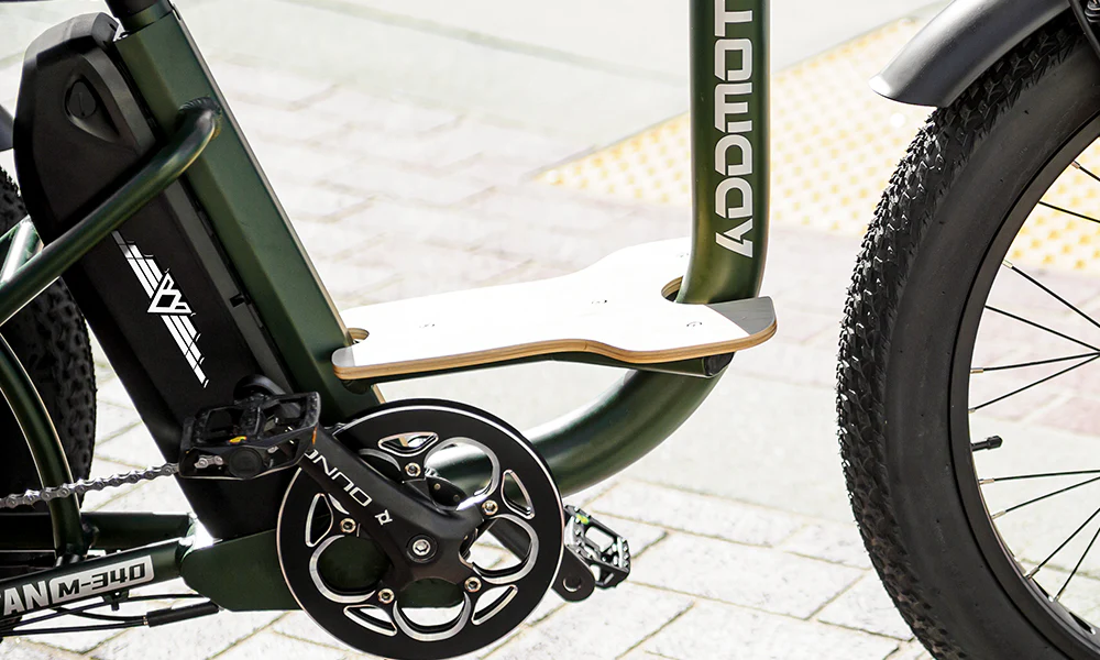 An electric trike for seniors featuring a low step frame for easy access and a sturdy wooden footrest board for added rider comfort, parked in a park setting surrounded by palm trees and neatly trimmed hedges.